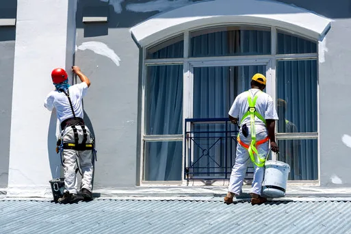 Two painters in safety gear working on a storefront facade with tall windows